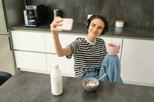 elegante joven mujer, estudiante tomando selfie en el cocina, Bebiendo té y comiendo desayuno, haciendo imágenes para social medios de comunicación aplicación foto