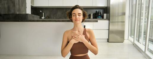 Heart chakra yoga. Portrait of woman practice mindfulness meditation at home, holding hands on chest with eyes closed and relaxed smile, sitting on rubber mat on kitchen floor photo