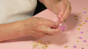 handgemaakt sieraden. wit Europese vrouw in wit t-shirt is maken handgemaakt bijouterie, kralen en armbanden Aan roze tafel Bij huis. ambachtsman werkplaats detailopname, statisch schot video