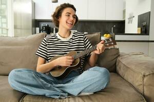 alegre joven mujer con ukelele, jugando musical instrumento, participación pequeño guitarra y cantando, sentado en sofá en cruzado piernas, descansando en vivo habitación foto