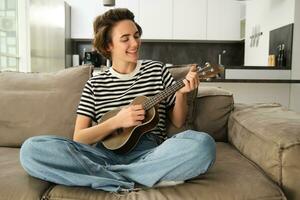 estilo de vida y aficiones concepto. joven sonriente mujer en sofá, jugando ukelele, canto y aprendizaje rasgueo modelo para favorito canción foto