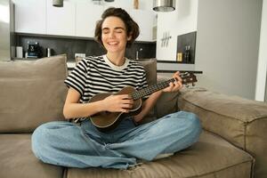 retrato de alegre joven mujer jugando su ukelele, canto y reír, sentado en vivo habitación a hogar foto