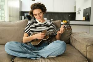 retrato de linda joven mujer sentado en sofá, aprende cómo a jugar ukelele, participación su música instrumento, cosecha acordes, descansando en vivo habitación foto