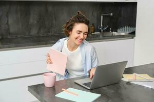 sonriente joven mujer reír, vídeo chats en computadora portátil, conecta a en línea trabajo reunión, estudios desde hogar, sostiene su computadora portátil, se sienta en cocina foto