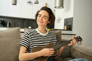 Close up portrait of beautiful smiling woman, playing ukulele, strumming strings with closed eyes and pleased face, sitting in living room photo