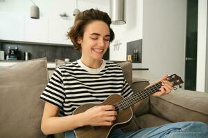 Portrait of young modern woman, musician playing ukulele at home, picking chords for new song, sitting on sofa in living room and smiling photo