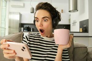 Portrait of woman binge watching her favourite tv show on smartphone, drinking tea and wearing headphones, gasping from shock, looking amazed at mobile screen photo