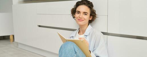 Portrait of creative young woman, artist sits on floor and draws in her planner, writes diary, smiling while doing art at home in cozy space photo