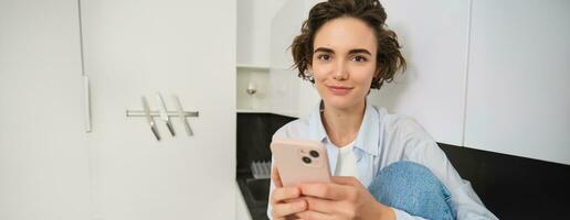 Smiling brunette girl sitting with smartphone at home, orders takeaway from mobile phone app, using online shopping application photo