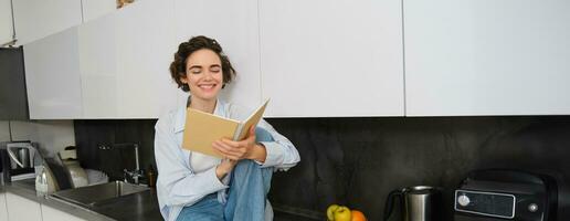 Lifestyle and people concept. Young woman enjoying weekend at home, reading notes in her journal, sits on kitchen counter with notebook and smiling photo