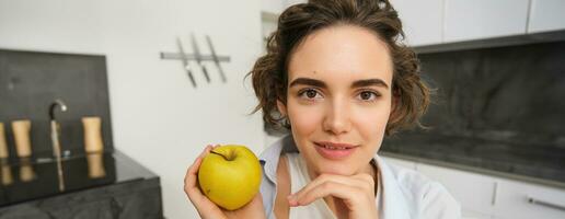 Close up portrait of young woman holding an apple, eating healthy fruits in her kitchen, smiling happily photo