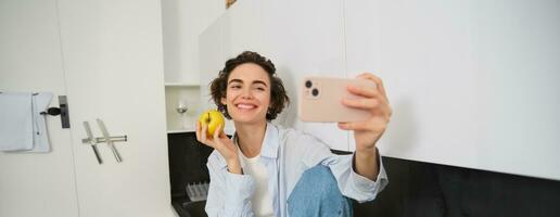 Beautiful girl blogger, takes selfie as she eats apple, shows how to lead healthy lifestyle to followers online, holds smartphone for photo