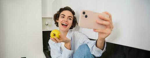 Beautiful woman eating an apple and recording herself on video. Cute girl with fruit, posing for photo, taking selfie in kitchen in happy mood photo