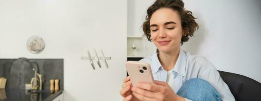 Smiling brunette girl sitting with smartphone at home, orders takeaway from mobile phone app, using online shopping application photo