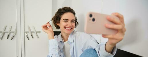 sonriente niña toma selfie en teléfono inteligente, posando para un foto en móvil teléfono aplicación, sentado en su cocina