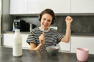 Carefree young woman, student in headphones, eating cereals for breakfast, listening music, singing and using soon as a microphone, having fun while having her meal in the morning photo