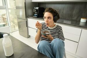 Image of stylish, modern woman, student eating cereals with milk for breakfast, holding bowl and spoon, listening music in wireless black headphones photo