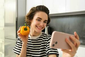 Smiling, beautiful young woman taking selfie with fresh yellow pepper, food blogger taking selfie with vegetables in the kitchen, vegan making a meal and recording video for social media photo