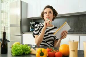 Portrait of thinking woman with notebook, cooking, writing down recipe ingredients, deciding on a meal for dinner, sitting near vegetables and chopping board in kitchen photo