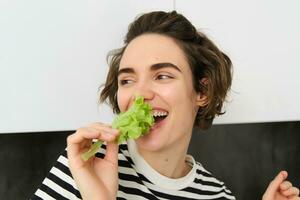 retrato de despreocupado vegetariano chica, comiendo verduras, mordedura lechuga hoja con contento sonriente rostro, teniendo sano bocadillo, gustos verduras, soportes en el cocina foto