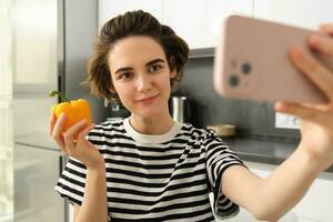 Close up portrait of cute brunette woman, taking selfie with yellow fresh pepper, recording video on smartphone, posing with vegetables in the kitchen, making cooking blog on social media app photo