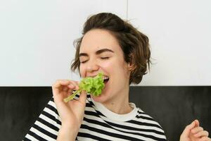 cerca arriba de gracioso linda mujer, vegetariano comiendo lechuga hoja y sonriente, concepto de sano dieta, niña gustos verduras, en pie en el cocina foto