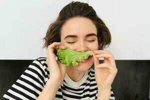 cerca arriba retrato de joven mujer, vegetariano chica, gustos comiendo verduras, posando con lechuga hoja y sonriente, posando en el cocina. concepto de sano comida y dieta foto