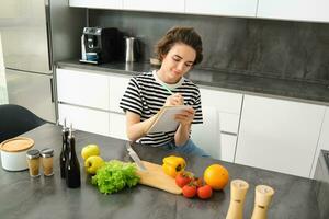 retrato de mujer escritura abajo su receta, haciendo notas en cuaderno durante cocinando, sentado en el cocina con el cortar tablero y verduras, preparando un comida foto