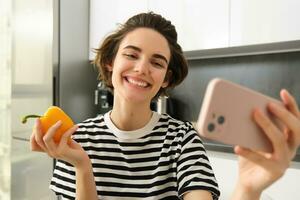Close up portrait of cute brunette woman, taking selfie with yellow fresh pepper, recording video on smartphone, posing with vegetables in the kitchen, making cooking blog on social media app photo