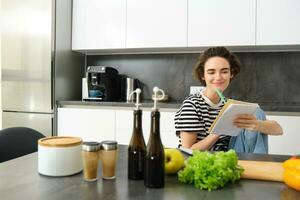 retrato de joven mujer mirando a Cocinando ingredientes en cocina mostrador y haciendo notas, escritura abajo recetas, pensando de comida para cena, preparando vegetariano comida foto