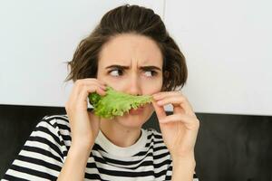 Portrait of woman frowning and grimacing, smelling fresh lettuce leaf with displeased face, standing in the kitchen, dislike vegetables photo