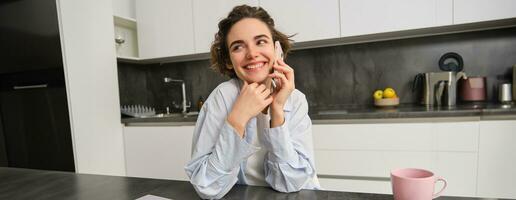 sonriente joven mujer negociaciones en móvil teléfono, llamadas alguien desde hogar, se sienta en cocina y tiene conversacion foto