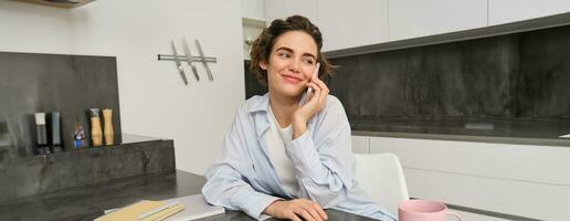 Cellular technology and people. Smiling brunette woman sits at home in kitchen, talks on mobile phone photo