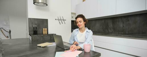 retrato de morena mujer lo hace tarea, trabajos desde hogar, prepara para examen en cocina, se sienta con cuaderno y ordenador portátil foto