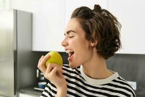 cerca arriba retrato de joven morena mujer mordiendo un manzana con placer, tiene satisfecho sonrisa en su rostro, en pie en el cocina, teniendo sano bocadillo para almuerzo, comiendo frutas foto