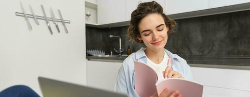 Young woman checking her schedule while working on remote from home, using laptop, looking at her daily planner, making notes, writing down information in notebook photo