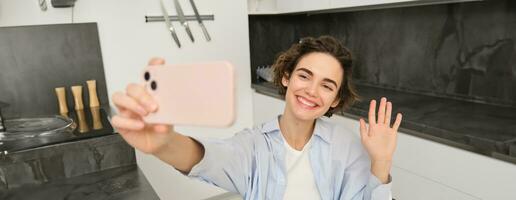 Happy, beautiful brunette girl takes selfie at home, poses for photo with smartphone