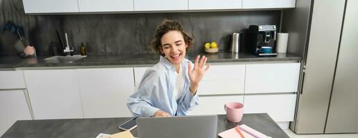 Happy young woman, tutor teacher students online. Girl connects to remote work meeting from her home laptop, says hello, waves hand at computer photo