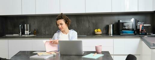 Portrait of working woman at home, checks her notebook, flips page, connects to online meeting via laptop. Girl learns programming remotely, studies course in internet photo