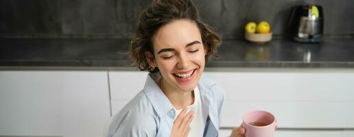 Happy mornings. Portrait of happy brunette woman, drinks cup of coffee in her kitchen and smiling, cozy and warm start of the day with cuppa photo