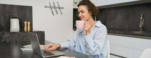Beautiful girl working from home, drinking coffee and looking at laptop. Young businesswoman sits in kitchen and manages her business via computer photo