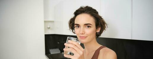 sano estilo de vida y hembra bienestar. joven hermosa mujer en pie en cocina y Bebiendo vaso de agua después rutina de ejercicio capacitación. aptitud niña corsé hidratado después gimnasio clase foto