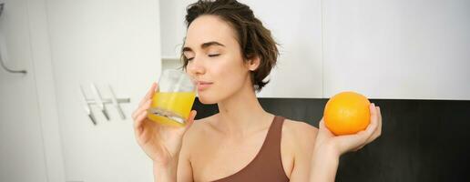 Image of sportswoman, fitness girl holding glass of juice and an orange, smiling, drinking vitamin beverage after workout, standing in her kitchen at home. Healthy lifestyle and sport concept photo