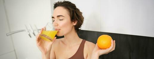 Healthy lifestyle and sport. Beautiful smiling woman, drinking fresh orange juice and holding fruit in her hand, enjoying her vitamin drink after workout at home, standing in kitchen photo