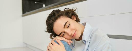 Close up portrait of beautiful, tender young woman smiling, looking at camera, sitting in cozy pose on kitchen floor, daydreaming photo