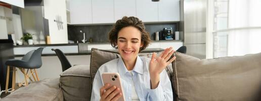 Lifestyle shot of happy girl talks on mobile phone, waves hand at smartphone camera, connects to online group chat on telephone app, sits on sofa at home photo