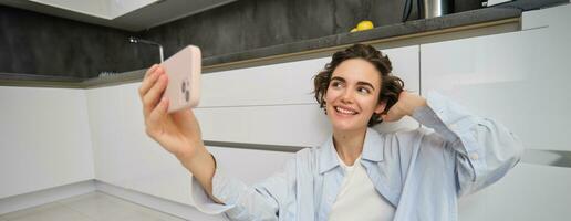 Portrait of young woman sits on kitchen floor with telephone, takes selfie on smartphone with app filters, poses for photo on mobile phone