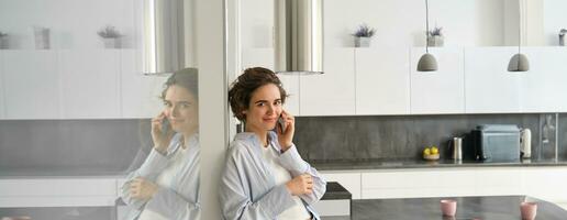 Beautiful woman talking on mobile phone, answers phone call and smiles, stands at home, leans to wall and chats photo