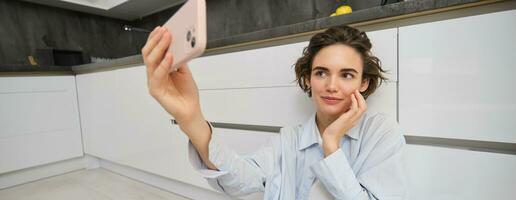 Portrait of young woman sits on kitchen floor with telephone, takes selfie on smartphone with app filters, poses for photo on mobile phone