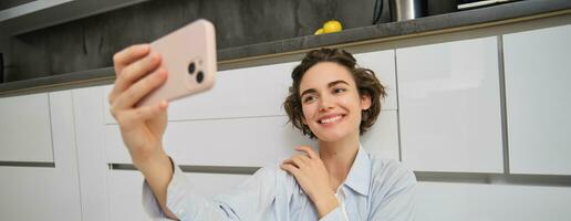 Portrait of young woman sits on kitchen floor with telephone, takes selfie on smartphone with app filters, poses for photo on mobile phone